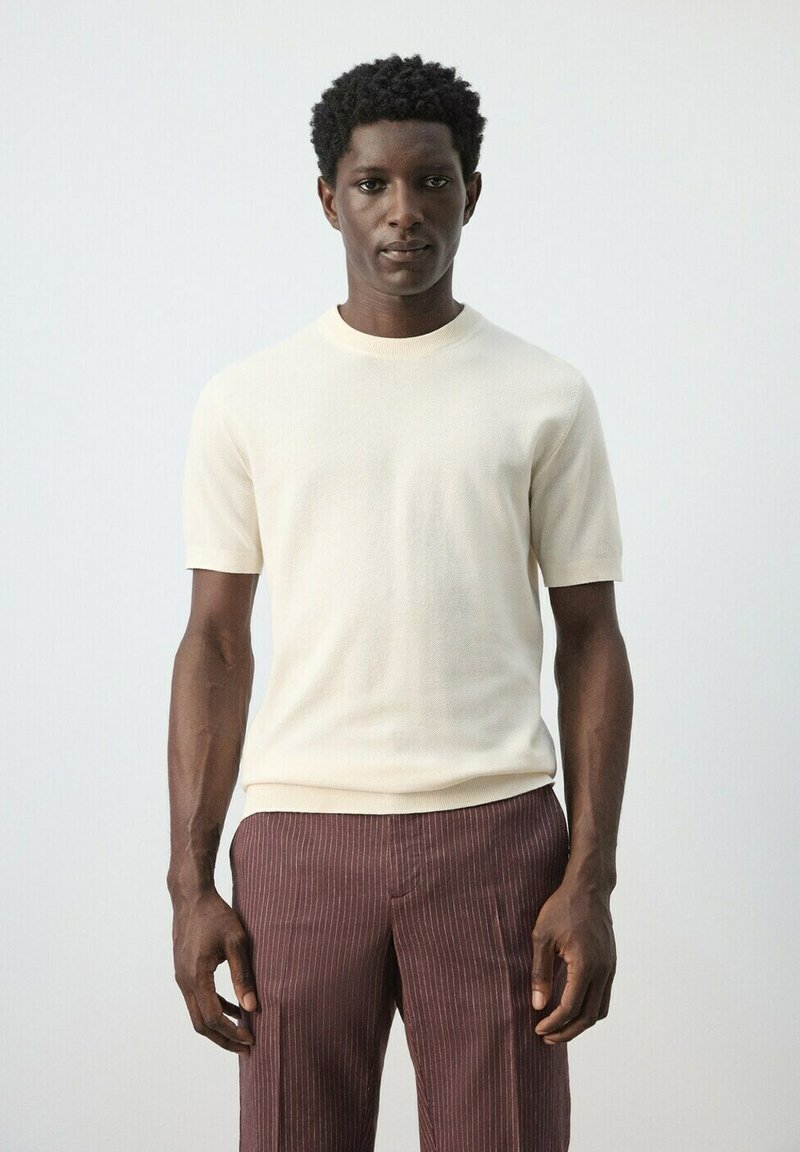 Mann steht und trägt einen cremefarbenen kurzärmligen Pullover und braune Nadelstreifenhosen vor einem schlichten hellgrauen Hintergrund.