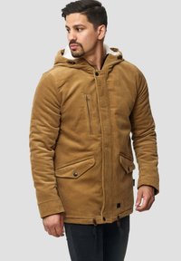 Hombre vestido con una chaqueta con capucha de pana color beige, con bolsillos de solapa abotonados y un bolsillo en el pecho con cremallera, mirando hacia un lado contra un fondo sencillo.