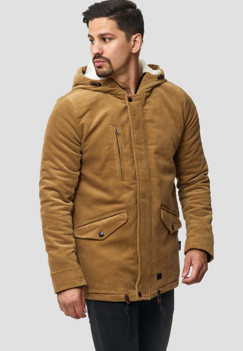 Hombre vestido con una chaqueta con capucha de pana color beige, con bolsillos de solapa abotonados y un bolsillo en el pecho con cremallera, mirando hacia un lado contra un fondo sencillo.