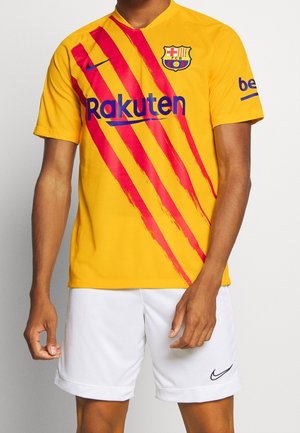 Hombre con camiseta de fútbol del FC Barcelona a rayas amarillas y rojas, pantalones cortos blancos de Nike, de pie contra un fondo liso.