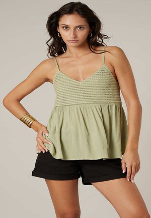 Vrouw draagt een mouwloos lichtgroen textuurtopje en zwarte shorts, poseert met hand op heup tegen een neutrale achtergrond.