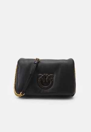 Sac bandoulière en cuir noir avec bandoulière chaîne dorée et logo doré en forme d’oiseau sur le devant, sur fond uni.