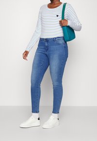 Camiseta de rayas de manga larga en azul y blanco con un pequeño logo de corazón, combinada con unos jeans ajustados azules y zapatillas blancas, sosteniendo un bolso color teal.