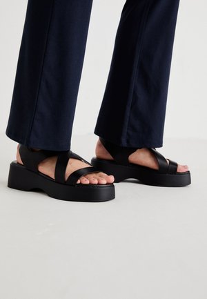 Feet wearing black platform sandals with wide straps, paired with dark navy straight-leg pants on a light floor.
