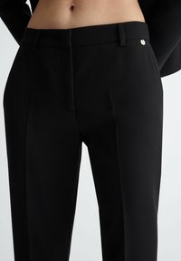 Pantaloni neri su misura realizzati in un tessuto liscio, con piega frontale e un piccolo dettaglio di bottone dorato in vita.