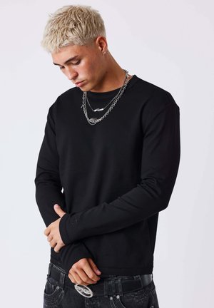 Jonge man met kort geblondeerd haar, draagt gelaagde zilveren kettingen, een zwart shirt met lange mouwen en donkere spijkerbroek, kijkt naar beneden met zijn handen gevouwen.