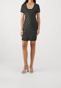 Svart klänning med korta ärmar som har ett diamantmönster och rundad halsringning. Bärs med vita strappy klackar, vilket visar en åtsittande silhuett.