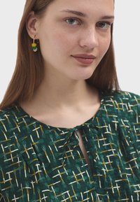 Zelená blůza s geometrickým vzorem a detaily na zavazování kolem krku. Obsahuje bílé a žluté mřížkové linie. Modelka nosí vícebarevné náušnice.