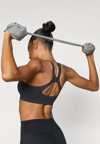 Μαύρο αθλητικό σουτιέν με σχεδιασμό με κοψίματα, συνδυασμένο με μαύρο κολάν με ψηλή μέση. Το μοντέλο κρατά μια λωρίδα αντίστασης φορώντας γκρι γάντια.