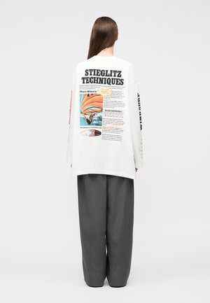 Person mit langen braunen Haaren steht mit dem Rücken zur Kamera, trägt ein weißes Langarmhemd mit bunten Windsurfing-Grafiken und dunkelgraue weite Hosen.