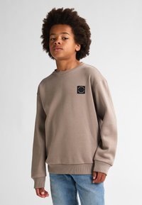 Un felpa marrone con colletto a girocollo e polsini a coste, caratterizzata da un piccolo logo nero ricamato sul petto. Indossata con jeans azzurri chiari.