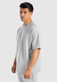 T-shirt grigio a maniche corte realizzato in tessuto morbido, con scollatura rotonda e un piccolo logo sul petto, esposto su uno sfondo neutro.