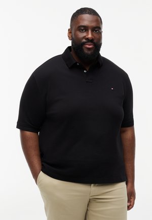 Mężczyzna z brodą w czarnej koszulce polo i beżowych spodniach, stojący z rękami w kieszeniach na białym tle.