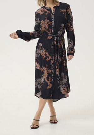 Femme portant une robe midi noire avec des motifs cachemire bruns et gris, manches longues, devant boutonné, ceinture à la taille, et sandales noires à brides.