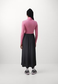 Persoon met lange gevlochten haren, gekleed in een roze turtleneck, zwarte midi-rok, witte sokken en zwart-witte sneakers, staand met de rug naar de camera gekeerd.