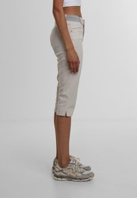 Helle beige Caprihosen mit schlankem Schnitt, mit einem grauen Taillenbund und Seitenschlitzen am Saum. Getragen mit grauen Turnschuhen.
