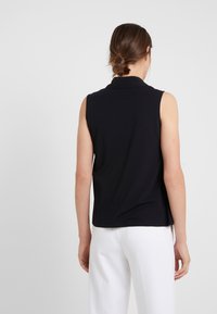 Mujer con cabello castaño recogido en un moño bajo, vestida con una blusa sin mangas negra y pantalones blancos, de pie de espaldas frente a un fondo sencillo.