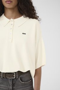 Krēmveida polo krekls ar apkakli, tam ir poga rinda un melns logotips. Kombinēts ar tumši zilām džinsa biksēm un brūnu jostu.