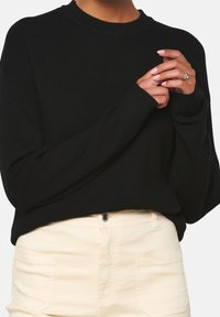 Mustat neulotut neuleet, joissa on pyöreä kaula-aukko ja pitkät hihat, rento malli ja ribbikuviointi, yhdistettynä vaaleisiin housuihin.