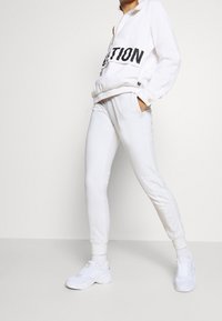 Biała kurtka na zamek błyskawiczny z czarnym napisem, elastycznymi mankietami oraz dopasowane białe spodnie dresowe. Oba wykonane z miękkiego materiału o delikatnym połysku.