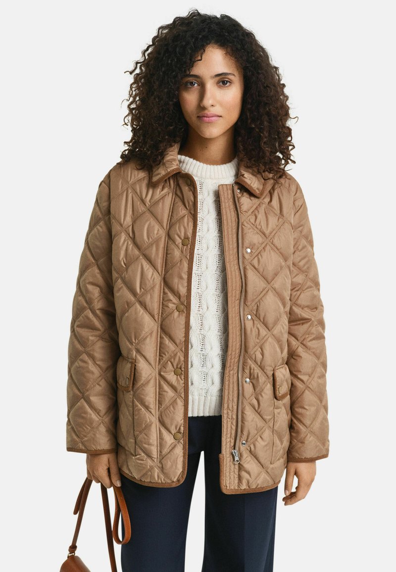 Bruine gewatteerde jacket met drukknopen en rits, zijzakken en geribbelde manchetten, gelaagd over een witte kabelgebreide trui.