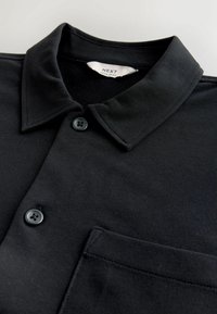 Polo negru dintr-un material texturat, având un guler clasic, două butoane negre și un buzunar pe piept pe partea stângă.