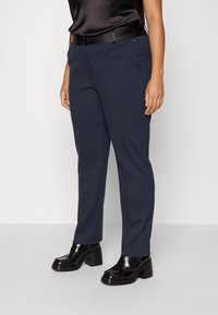 Marineblaue, maßgeschneiderte Hosen mit glatter Textur, ausgestattet mit Seitentaschen, Gürtelschlaufen und schlanker Passform. Kombiniert mit glänzenden, schwarzen Absatzschuhen.