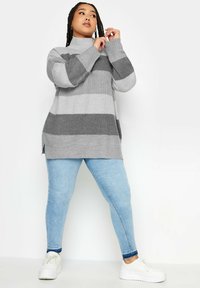 Grau-weiß gestreifter Strickpullover mit hohem Kragen, der über eine gerippte Textur, seitliche Schlitze verfügt und mit hellblauen Jeans und weißen Turnschuhen kombiniert ist.