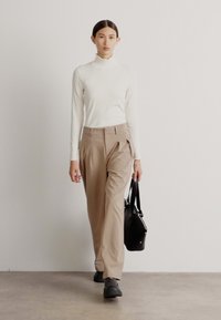 Beige vide bukser kombineret med en hvid turtleneck. Modellen bærer en sort taske. Gulvet er i lys farve, og baggrunden er minimalistisk.