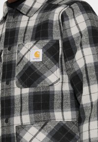 Grau und schwarzes kariertes Flanellhemd mit Druckknopfverschluss, ausgestattet mit einer linken Brusttasche und einem Carhartt-Logopatch auf der Tasche.