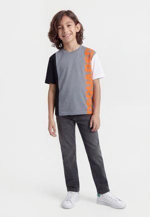 Usmievavý chlapec s vlasmi po plecia stojí oblečený v tričku s blokmi farieb sivá, čierna a biela s oranžovým nápisom, sivých džínsoch a bielych teniskách.