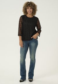 Top in pizzo nero con maniche a tre quarti su un sottogiacca uniforme, abbinato a jeans chiari a zampa d'elefante e scarpe nere con punta.
