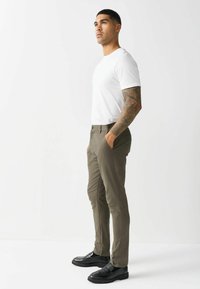 White fitted t-shirt paired with olive green trousers. Sleek black shoes with a textured finish. Model posed sideways, hands in pockets.