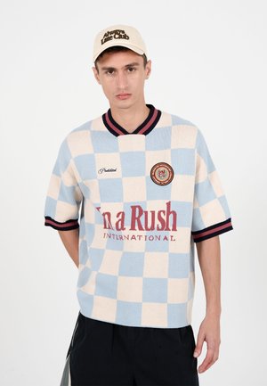 Jeune homme portant une casquette beige avec le texte « Always Late Club », une chemise à manches courtes à carreaux bleus et crème avec un texte rouge, et un short noir sur fond blanc.