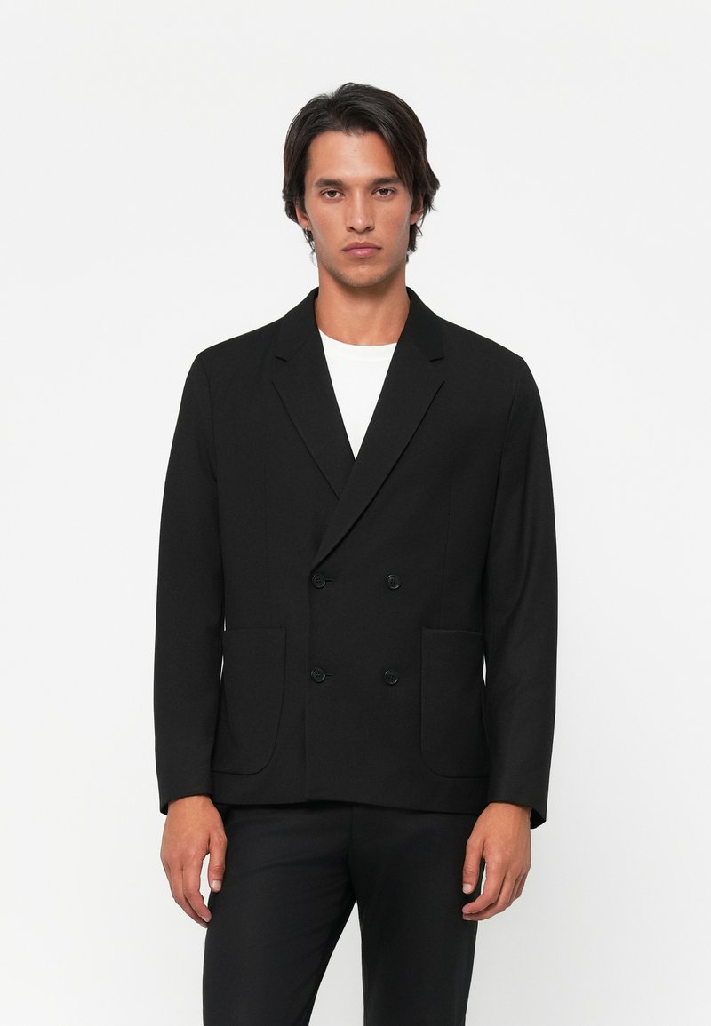 Blazer negro de doble botonadura con dos bolsillos frontales, solapas muescas y una textura suave, usado sobre una camisa blanca. Ajuste entallado.
