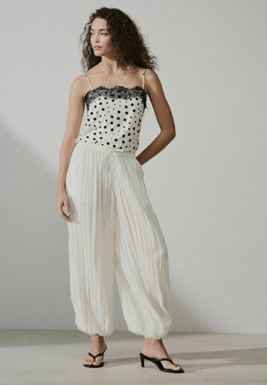 Femeie cu părul creț, purtând un maiou alb cu buline și pantaloni albi largi plisati, stând cu o mână în buzunar și privirea îndreptată spre stânga.