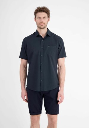 Man draagt een donker kort mouw knoophemd en donkere opgerolde shorts, staand tegen een effen lichte achtergrond.