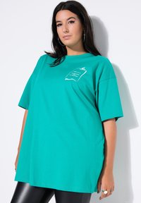 Turkis oversized t-shirt lavet af bomuld, med korte ærmer og et hvidt grafisk print på venstre bryst. Glat tekstur.