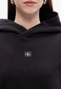 Felpa nera con cappuccio, realizzata in un tessuto texturizzato e con una patch quadrata del logo che presenta la scritta "Calvin Klein" e una "K" stilizzata stampata.