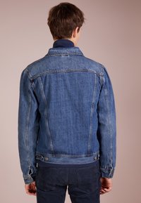 Mladý muž v modré džínové bundě a tmavých kalhotách, stojící zády k prostému béžovému pozadí.