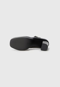 Black high-heeled shoe with a smooth finish and a textured rubber sole. Features a rectangular heel and a decorative accent on the upper.