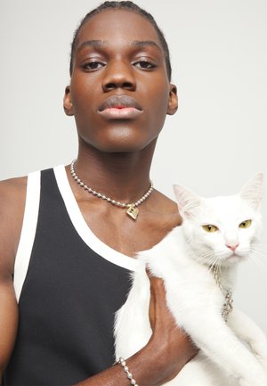 Persona joven con una camiseta sin mangas en blanco y negro y un collar de perlas sostiene un gato blanco con ojos amarillos contra un fondo liso.