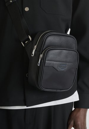 Borsa a tracolla nera con trama, dotata di due scomparti con cerniera, indossata sopra abbigliamento nero con orlo di camicia bianca visibile sotto.