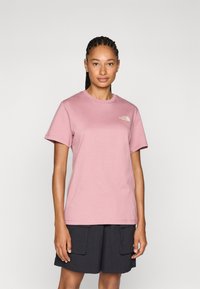 Rosa bomullströja med korta ärmar, rund halsringning och en liten vit logotyp på vänster bröst. Modellen bär mörka cargoshorts.