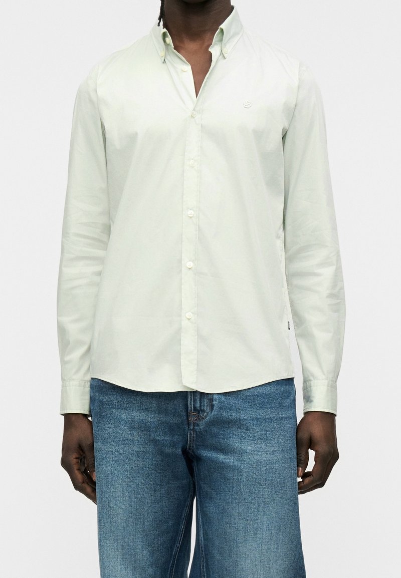 Homme portant une chemise à manches longues vert clair boutonnée et un jean bleu, debout devant un fond blanc uni.
