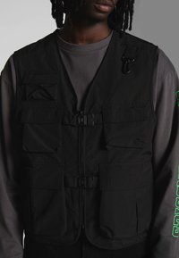 Schwarze Utility-Weste mit mehreren vorderen Taschen, einem zentralen Reißverschluss, Schnallen und einem verstellbaren Riemen; getragen über einem dunkelgrauen Langarmshirt.