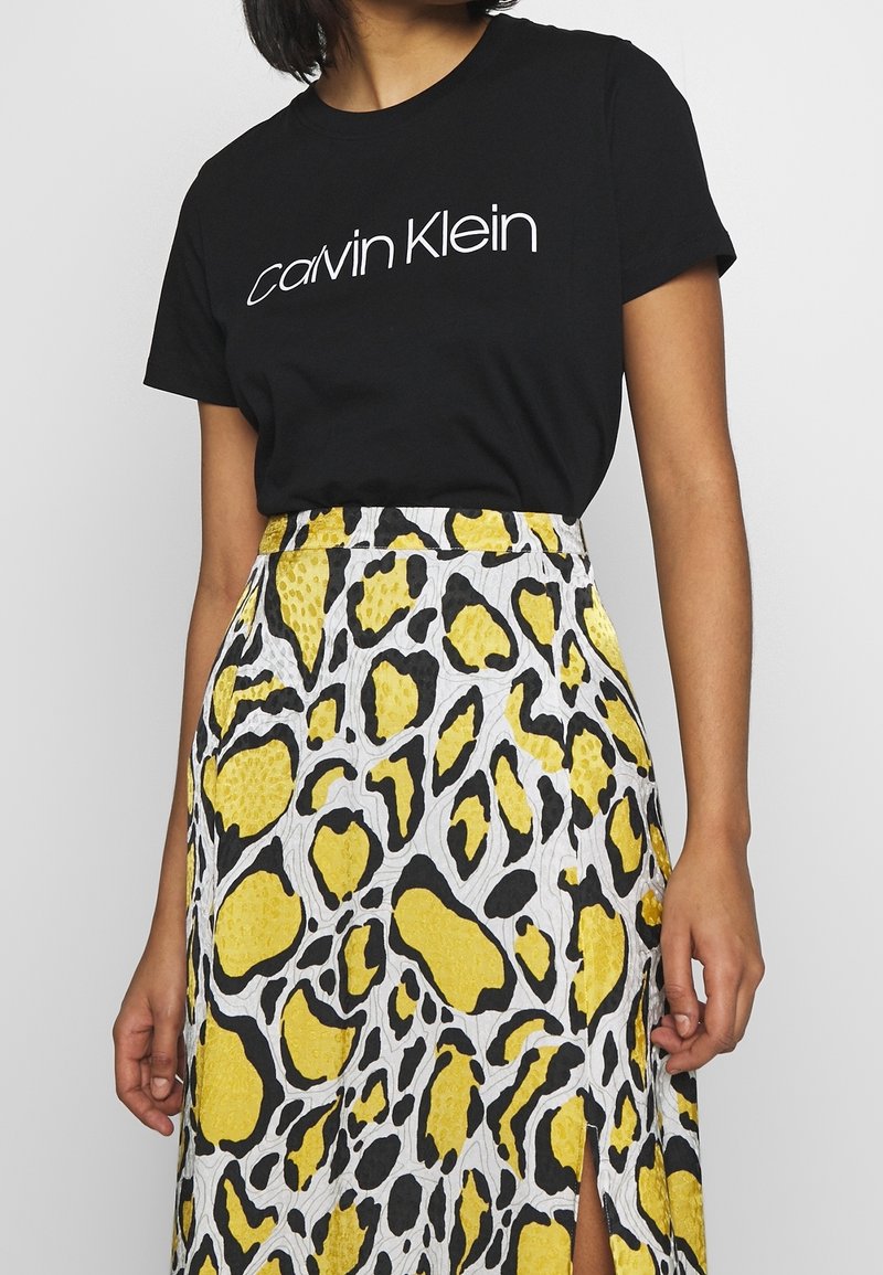 Černé bavlněné tričko s bílým logem „Calvin Klein“, doplněné žlutou a černou vzorovanou sukní s rozparkem a texturovanými detaily.