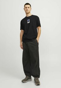 T-shirt nera in cotone con patch del logo, abbinata a pantaloni grigi scuri a vestibilità ampia e sneakers grigie. Design semplice e silhouetto rilassato.