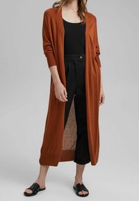 Cardigan long à devant ouvert de couleur orange brûlé, en maille avec des poignets côtelés, porté sur un haut noir et assorti à un pantalon noir court et des sandales.