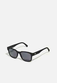 Ochelari de soare negri cu un finisaj mat, rame pătrate, lentile gri închis și un accent logo verde pe lateral. Design texturat al bratelor.